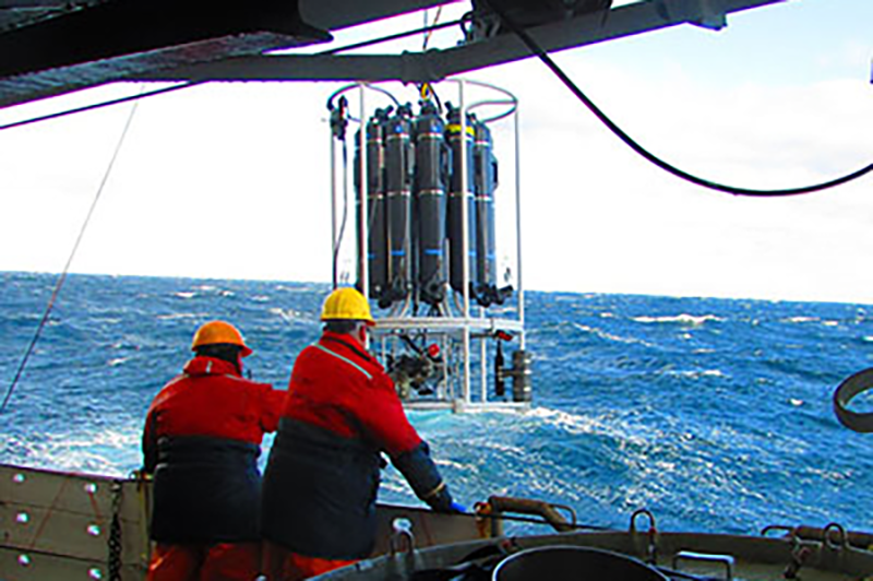 two men on deck with a large piece of equipemnt hanging fro ma wire over the side of the ship