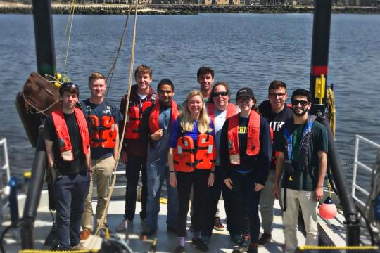 Ten people wearing orange life vests stand at the back end of a research vessel with a large, metal, black arch above them. The vessel is on the water with several buildings on the shore in the background.