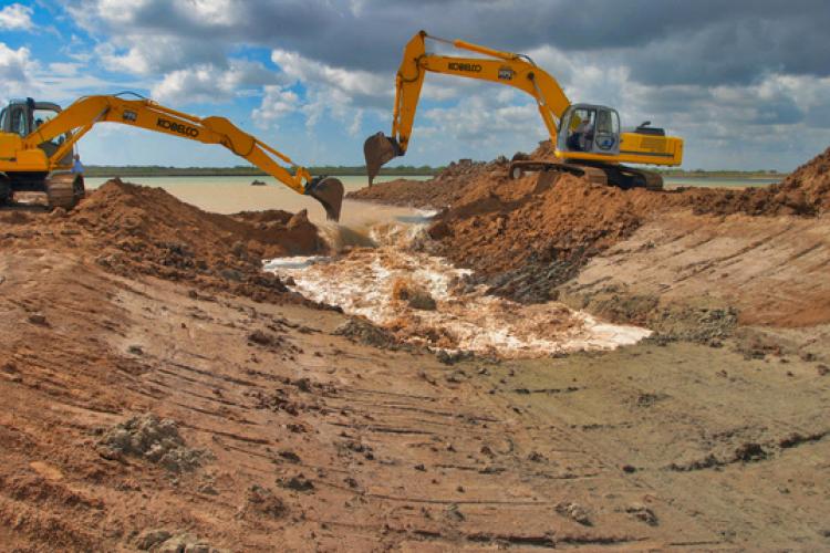 dam-migration-miss/md_077astIK2bCc.jpg Wide shot image showing two orange excavator machines digging up orange brown dirt and water coming in from the background